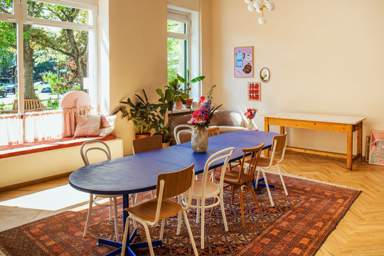 Lichtdurchfluteter Raum mit großer Fensterfront, blauer Tisch in der Mitte des Raumes mit acht Stühlen auf rotem Teppich, Blumenvase auf dem Tisch, Pflanzen im Raum, moderne Bilder an beige gestrichener Wand