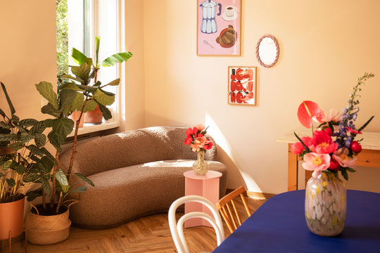 Detailansicht von lichtdurchfluteten Raum von Feasts of Eden mit grauer Couch, kleinem pinken Couchtisch, Pflanzen, modernen Bildern an der Wand und blauem Tisch mit Glasvase im Vordergrund